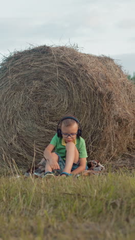 niño pequeño con camisa verde, auriculares y gafas, descansa la cabeza sobre la rodilla, la mano sobre la cabeza, centrándose en la música, en el fondo de una amplia tierra de cultivo