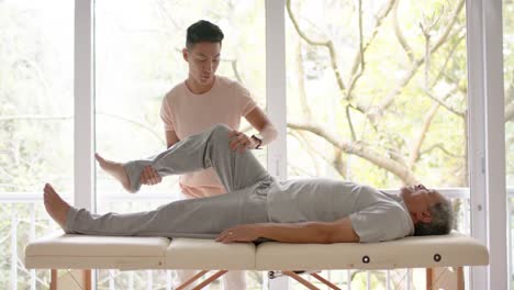 diverse male physiotherapist advising and senior male patient exercising on couch, in slow motion