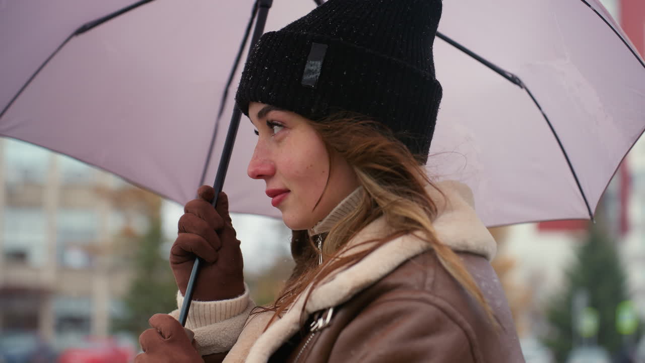Cheerful young woman in black knit cap and brown shearling jacket holding umbrella while walking slowly on cold overcast day, snowflakes on her hat, enjoying quiet moment outdoors in urban winter