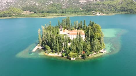 Aerial-shot-of-Visovac-Monastery-amidst-trees,-drone-flying-over-Christian-building-near-mountain-against-sky---Krka,-Croatia