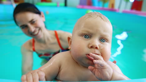 lovely caucasian baby with big blue eyes chewing fingers looking at camera. sweet kid is held by the coach in the swimming pool.