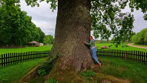 Young-Caucasian-woman-embrace-oak-rugged-trunk,-wonderful-green-scenery