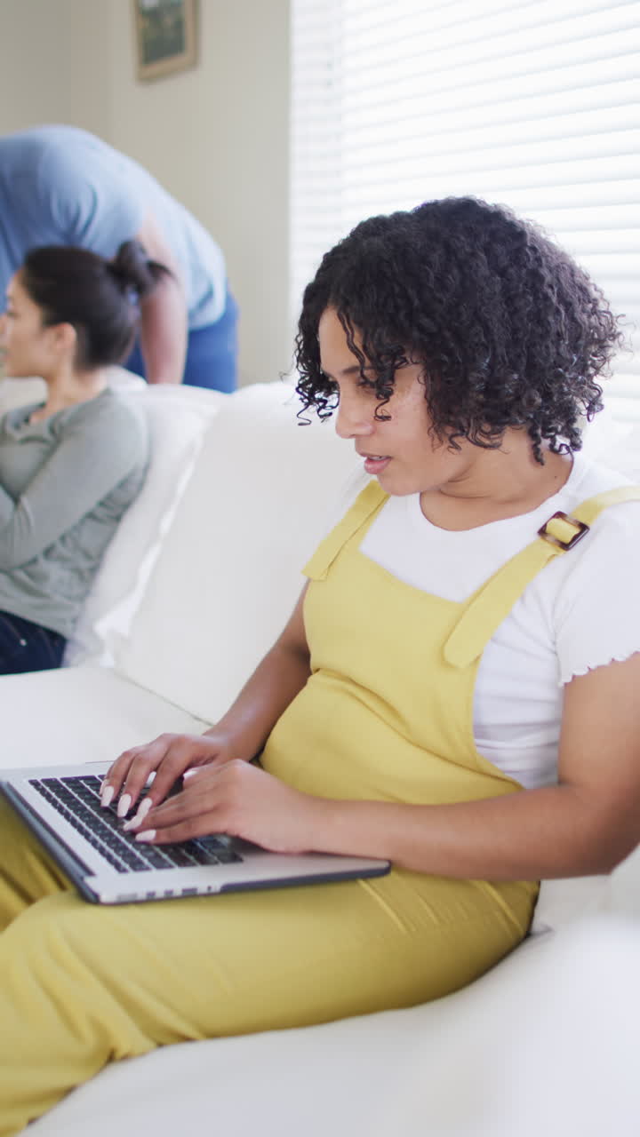 Vertical video of biracial woman on couch using laptop, with friends in background, in slow motion