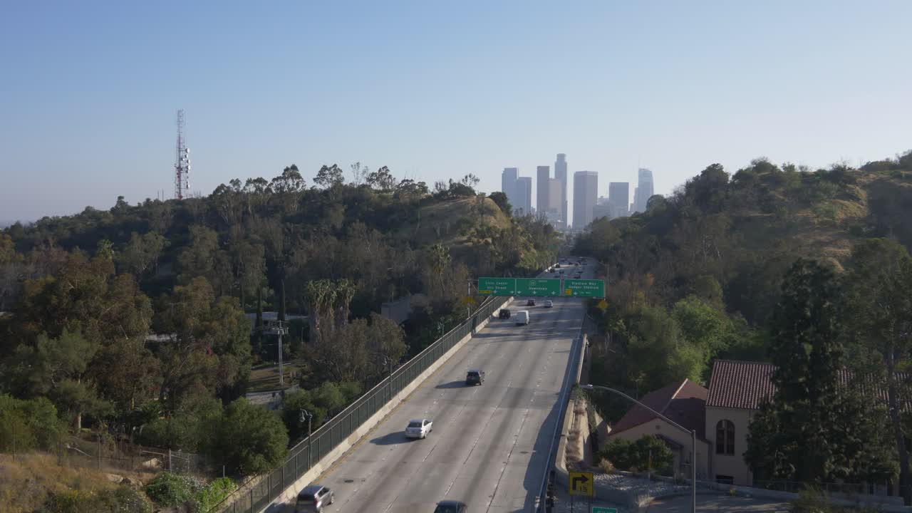 Drone view of a scenic highway winding through hillside greenery into downtown Los Angeles. A red-tiled landmark anchors the frame, blending natural contours with urban skyline energy