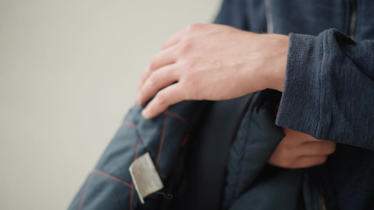 Primer plano de una persona sosteniendo y ajustando el cuello de una chaqueta acolchada azul con costuras naranjas, vistiendo una sudadera con capucha oscura con cremallera. La mano sujeta la tela cerca de los tiradores metálicos de la cremallera mientras el otro brazo descansa.