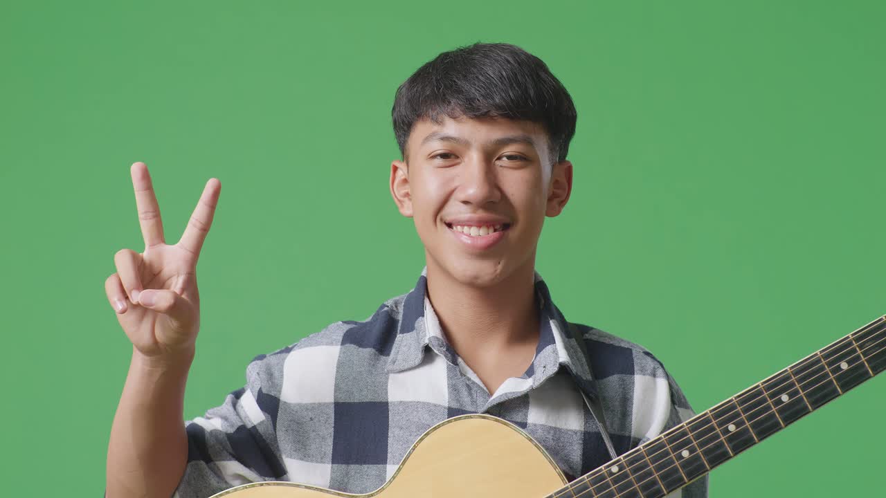 기타를 들고 있는 아시아 청소년의 클로즈업은 녹색 스크린 배경에서 평화와 미소를 지는 몸짓을 보여준다.