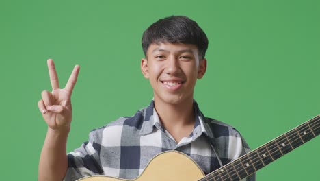 primer plano de un adolescente asiático con una guitarra mostrando un gesto de paz y sonrisa en el fondo de la pantalla verde
