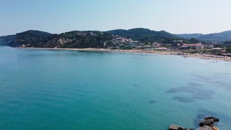 Greek-island-bay,-aerial-pan-left-view-of-people-swimming-and-sunbathing-on-beach-in-San-Stefanos-Corfu