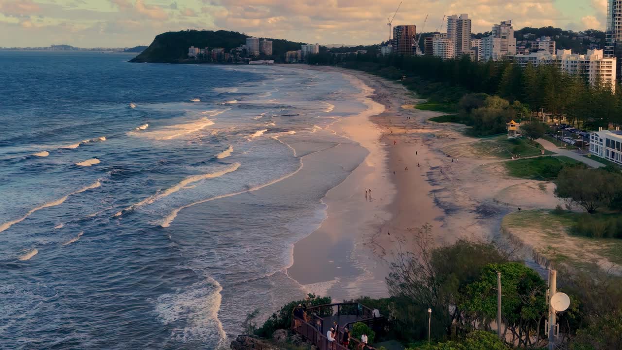 Aerial view of a sandy beach with gentle waves and a distant city skyline under a colorful sky.