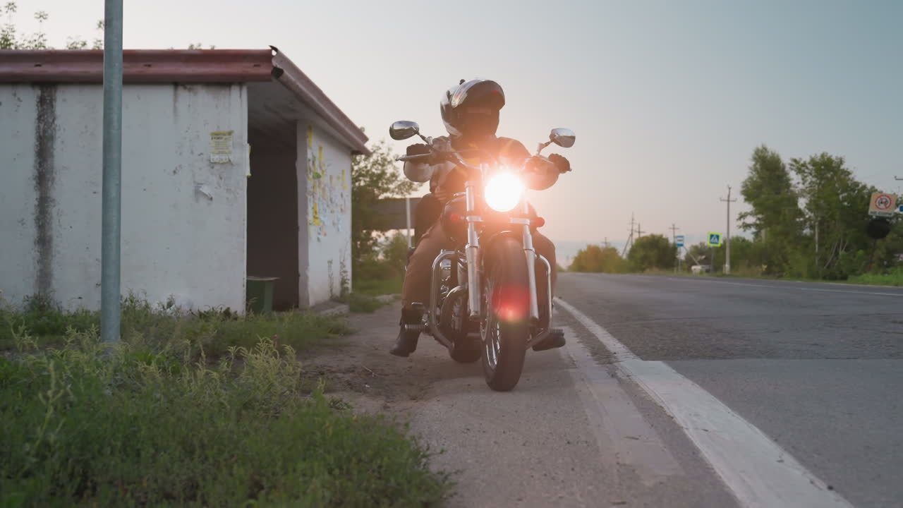 Motorbike rider in helmet drives off from roadside with bright headlight beaming, worn bus stop shelter beside, distant car parked on opposite lane, evening sky and long stretch of road