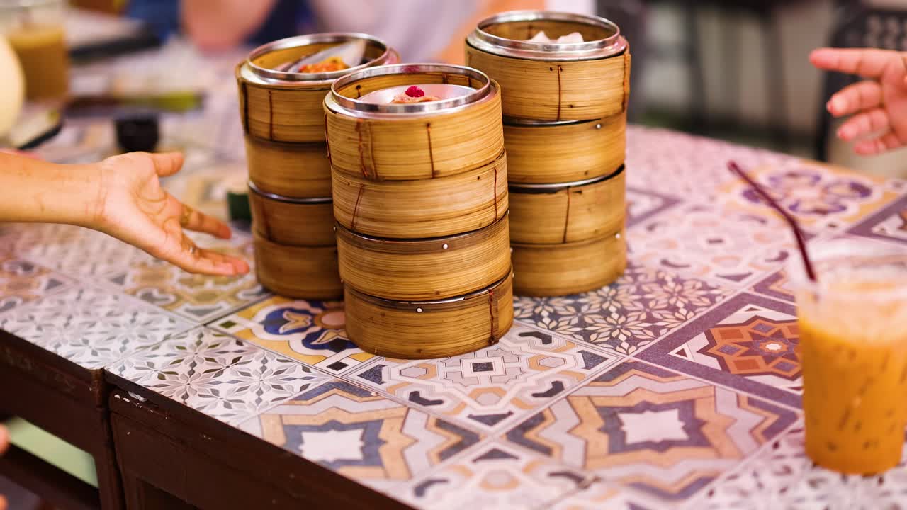 Hands reach for bamboo steamers filled with dim sum on a patterned table, creating a lively dining scene