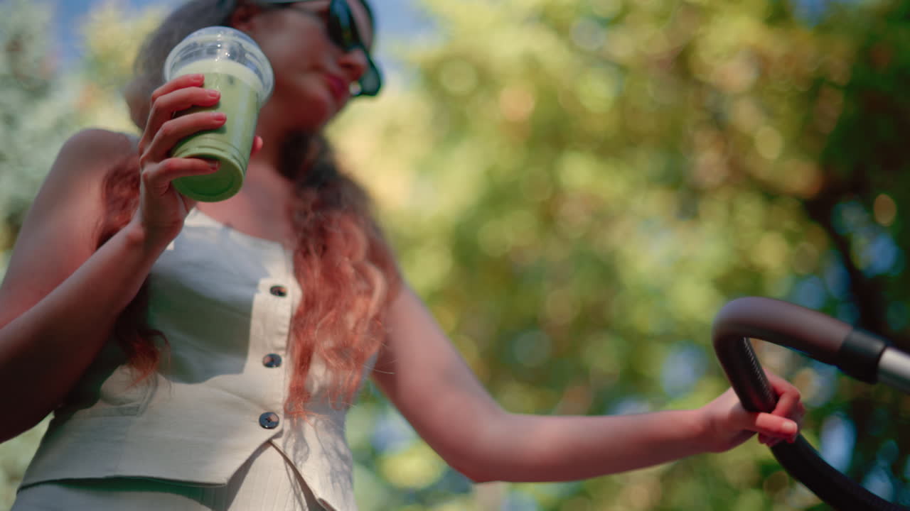 Close up of woman holding an iced matcha latte outdoors while pushing a stroller