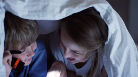 close-up view of cute little boy and her sister using a flashlight talking and staring at something under the blanket at night