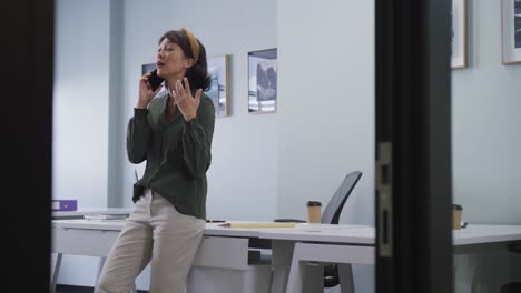 biracial businesswoman standing at table and talking on smartphone alone at office