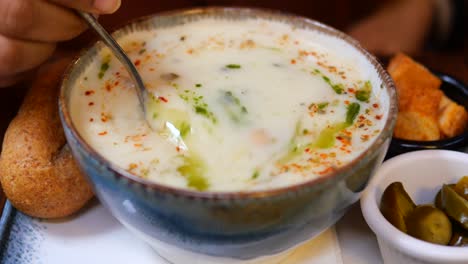 a bowl of creamy soup with chickpeas, bread, and pickles