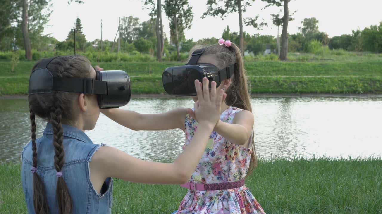 dos niñas felices juegan en gafas de realidad virtual, sentadas en la hierba del parque, con el telón de fondo de un hermoso río al atardecer en un día de verano.