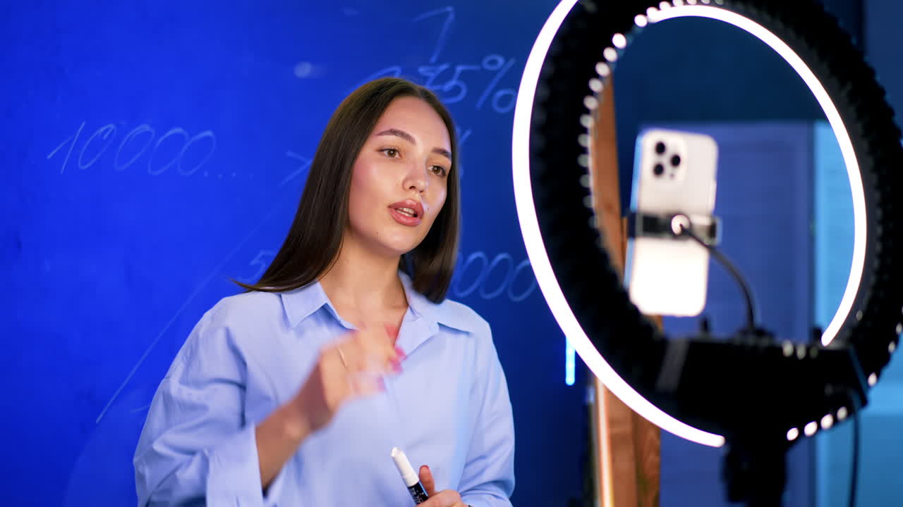 Attractive Caucasian lady with long dark hair wearing blue shirt. Woman taking video on her phone with light ring. Streaming concept.