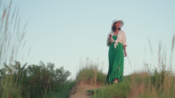 Avventuriera che scende una collina erbosa con in mano un bicchiere di vino, che indossa un cappello da sole e un abito verde fluente, con uno sfondo di fiume distante, un cielo al tramonto splendente, serena esplorazione all'aperto