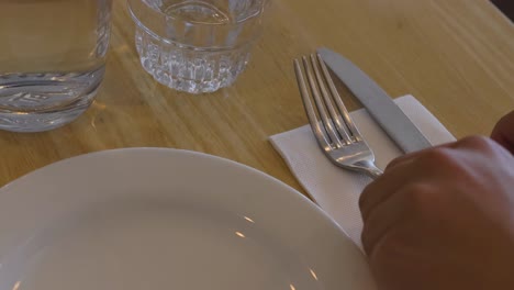 a person organizes cutlery and a glass beside a plate on a wooden table.