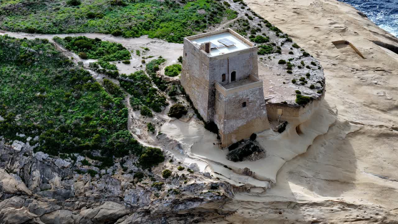 Scatto aereo dall'alto della torre di Xlendi sull'isola di Gozo a Malta durante una giornata estiva luminosa e soleggiata, con un'antica architettura in pietra calcarea che contrasta con i vivaci arbusti mediterranei verdi e le rocce dorate