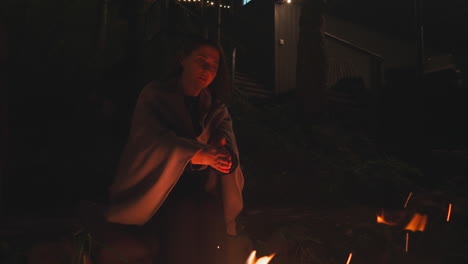 woman gazes at bonfire keeping hands above flame. young woman perches on stones next to fire pit in darkness near camping house. woman in warm blanket