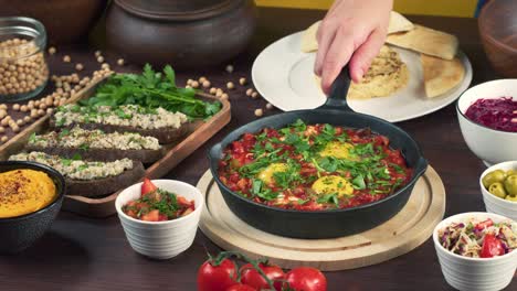 traditional israeli cuisine. shakshuka in pan decorated with parsley, forshmak and hummus on table. national jewish dishes. eggs fried in tomato sauce, dip, pita, middle eastern culture