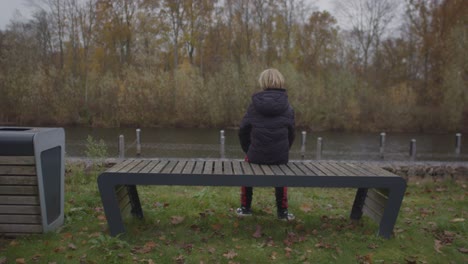 adolescente sentado en un banco del parque con vistas al río en otoño