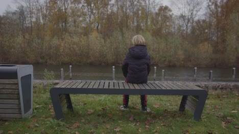 teenage boy sitting down on park bench overlooking river in autumn