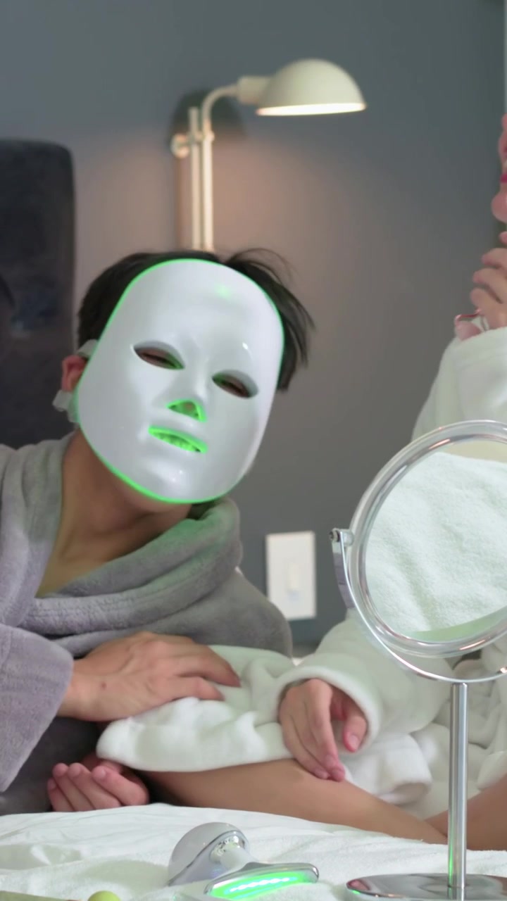 Video vertikal: Meraih perangkat, wanita memasangkan masker LED pada pria di tempat tidur yang mengenakan jubah untuk perawatan kulit.