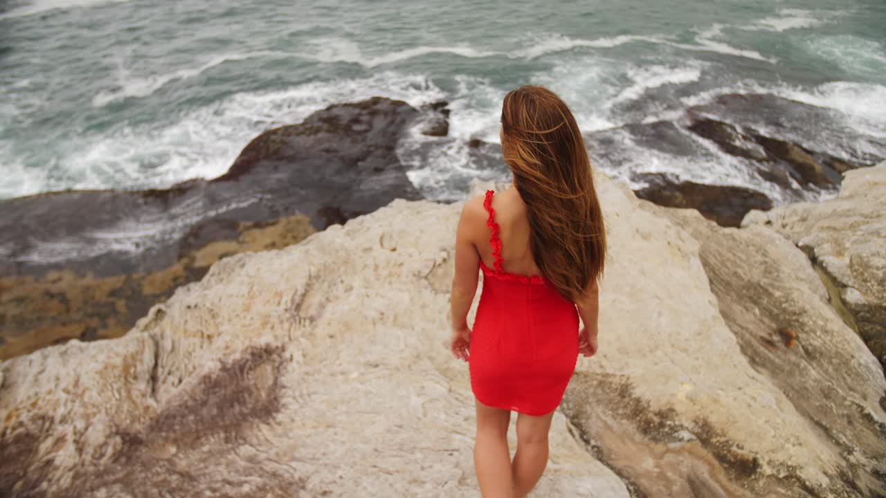 빨간 드레스를 입은 섹시한 소녀가 바위 위에 서서 파도를 배경으로 하늘을 올려다본다 - 동부 교외, 시드니, 뉴사우스웨일즈, 호주의 바위가 많은 해변 해안