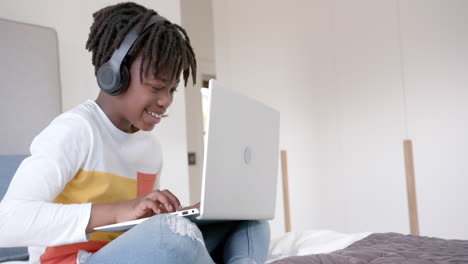 happy african american boy wearing headphones sitting on bed using laptop at home, slow motion