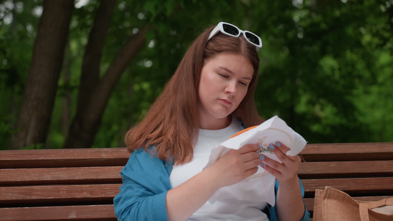 Young seamstress sitting on wooden park bench embroidering white fabric with needle, wearing white outfit and blue shirt, focused on delicate hand stitching while enjoying peaceful summer atmosphere