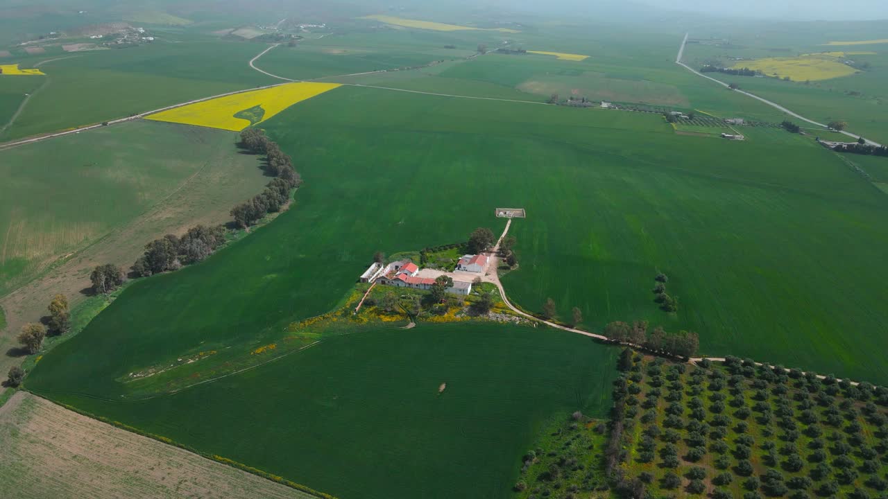 Aerial view of a green field with a big red farm and yellow flowers, there are green fields and mountains in the distance.