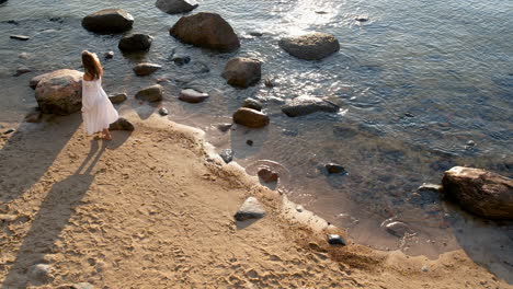 vista aérea de arriba hacia abajo de una mujer joven en un vestido de sol blanco caminando por la playa de arena con rocas en el agua del mar al amanecer