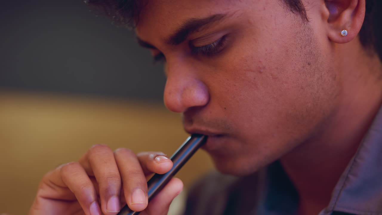 Extreme close-up of young man sipping from black straw with thoughtful expression, fingers gently holding straw, soft indoor lighting highlighting facial texture and subtle emotion, suggesting quiet focus or contemplation