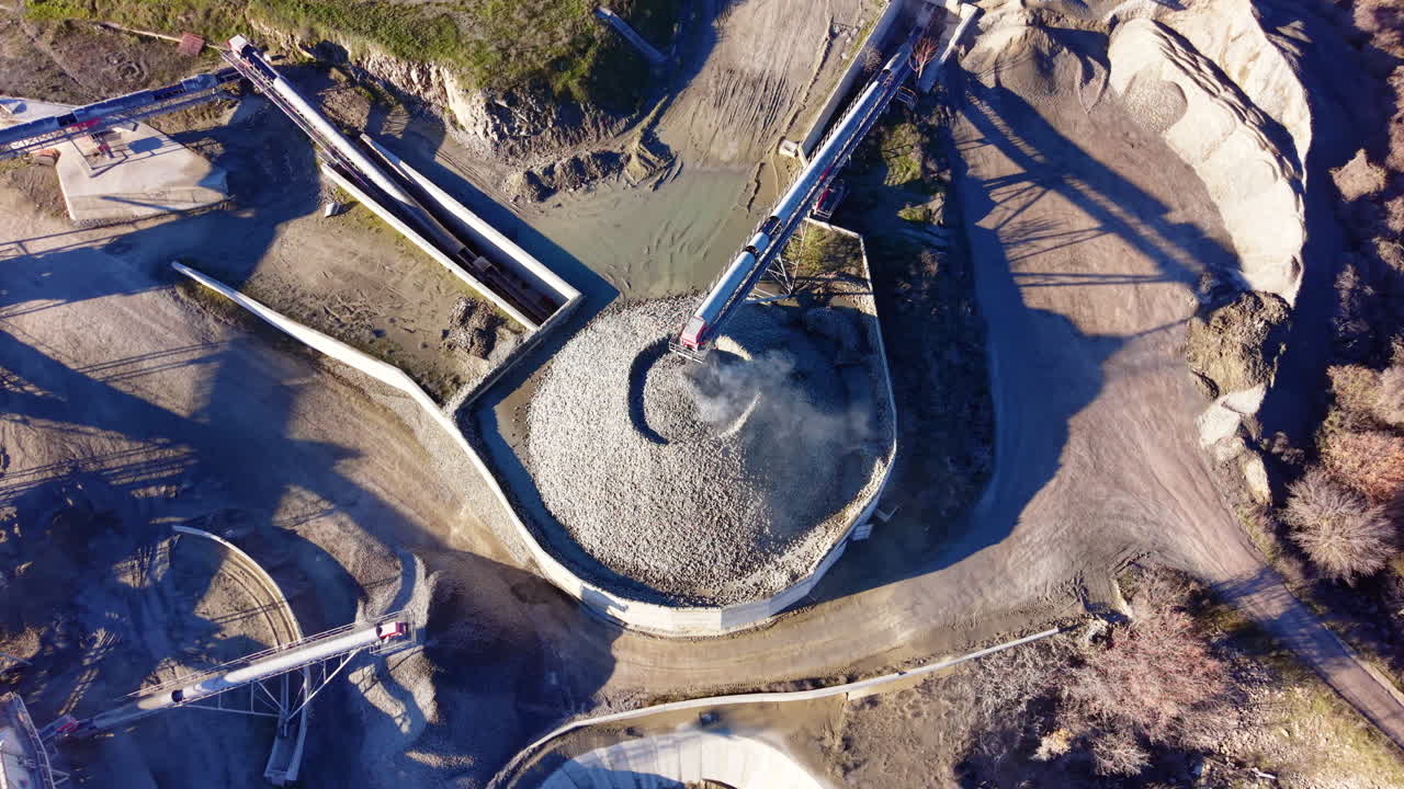 Large circular pile of crushed stone being fed by a long conveyor belt in a processing plant. Aerial top view of circular stone stockpile and conveyor system
