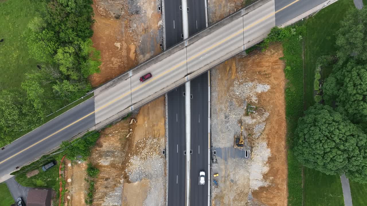 Aerial top down of American overpass crossing a highway under construction, surrounded by greenery and red soil. Ideal for themes like infrastructure, travel, or rural development. PA, United States.