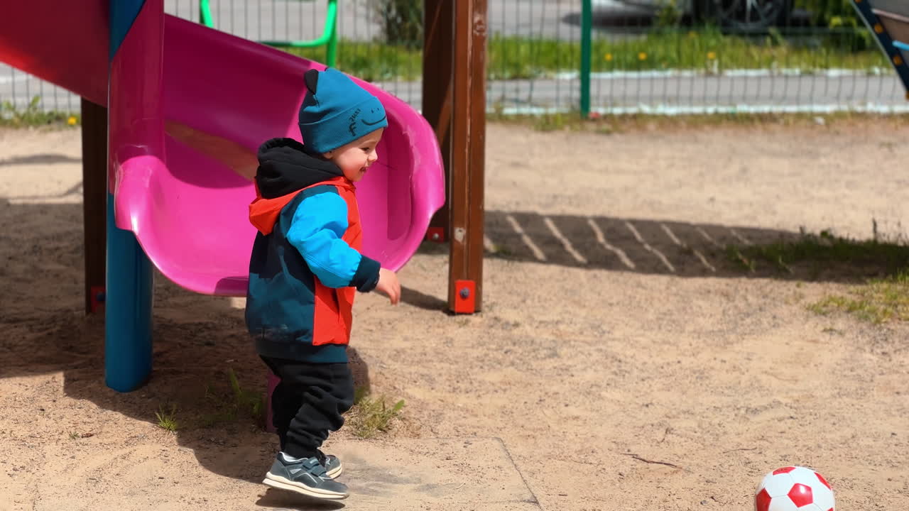 Little toddler tries to climb on the slide. Happy smiling boy grabs the ball and runs, falls, stands up and runs again.