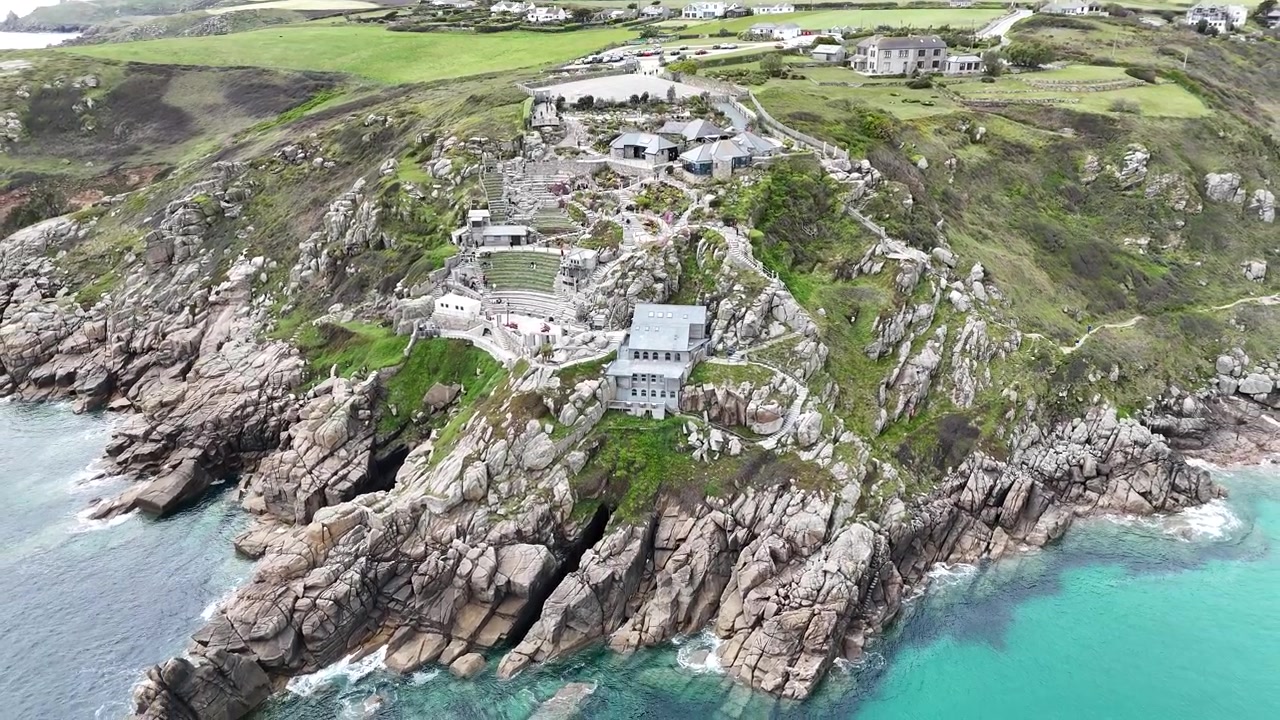 Rivelazione aerea con drone che cattura lo spettacolare Minack Theatre, arroccato in alto sopra coste rocciose e vivaci acque turchesi lungo la costa della Cornovaglia.