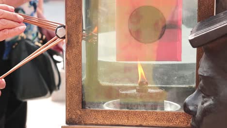 close-up of hands placing incense sticks into a flame behind a glass enclosure.