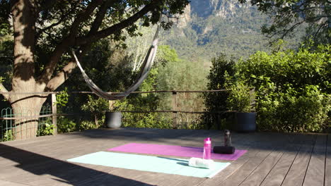 Dos-Esteras-De-Yoga,-Botellas-De-Agua-Y-Toallas-En-Cubierta-En-Un-Jardín-Soleado,-Espacio-Para-Copiar,-Cámara-Lenta