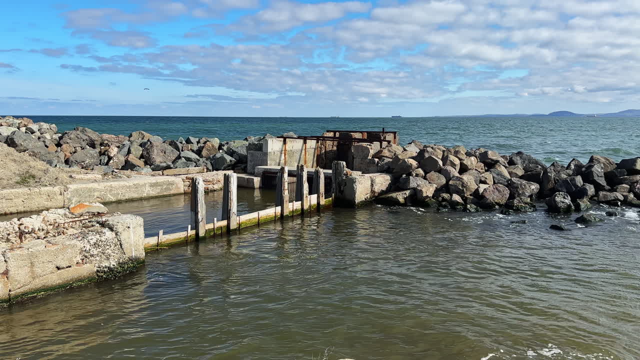 Udsigt til en forfalden anløbsbro og klipper langs kystlinjen med havet og skyer i baggrunden i dagtimerne.