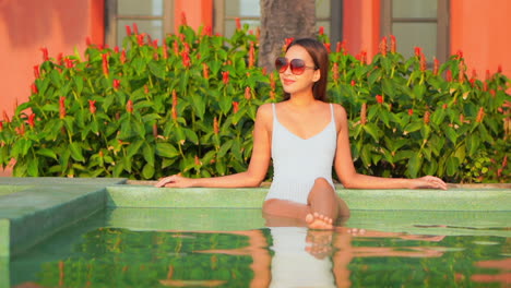 mujer sexy en traje de baño blanco sentada en el agua de la piscina poco profunda cruzó las piernas apoyándose en las manos en el salón del hotel tropical en miami - durante el día, cámara lenta