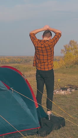 young man in red checkered shirt goes out of blue tent on meadow in early morning slow motion backside view