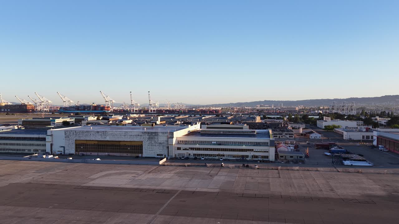 Amplia vista aérea con un paneo lento sobre la antigua estación aeronaval en Alameda, California, revelando el paisaje industrial y la concurrida terminal de contenedores del puerto de Oakland al fondo.