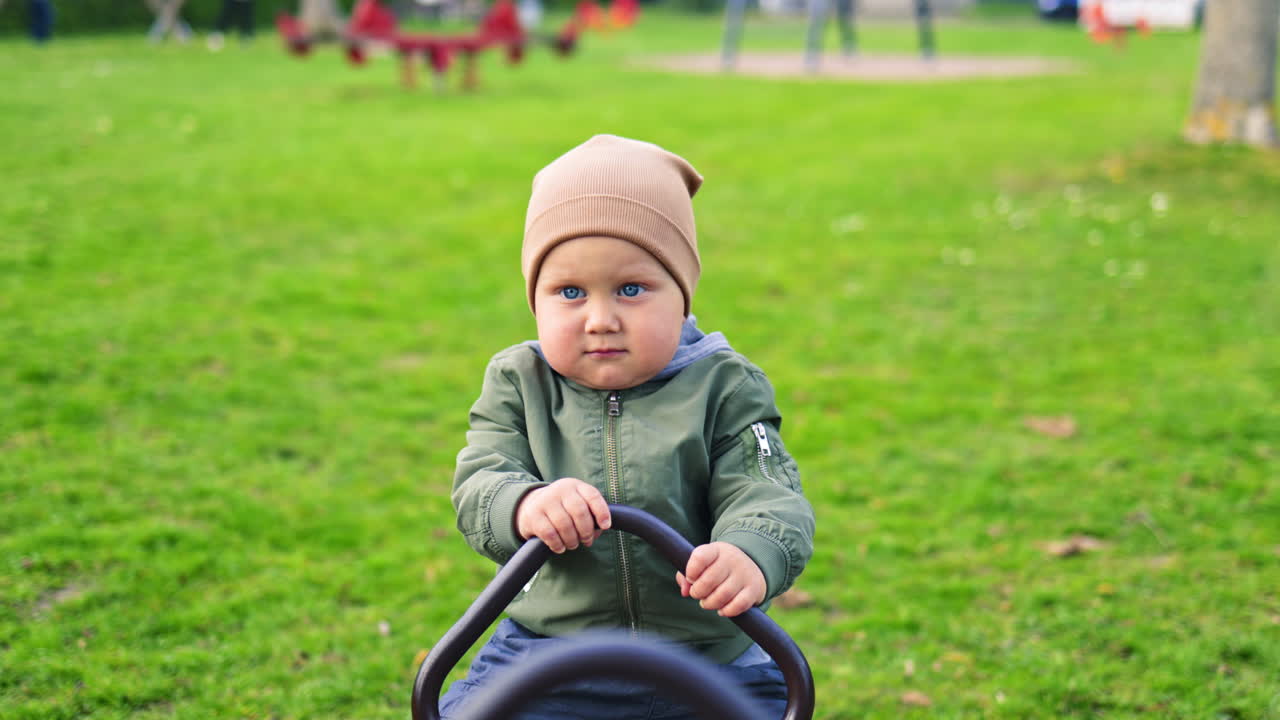 Un bambino piccolo va sull'altalena in un parco giochi. Un bambino di 2 anni con giacca e pantaloni cammina nel parco all'aperto. Stagione primaverile. Infanzia. Un bambino felice e allegro fa i primi passi.