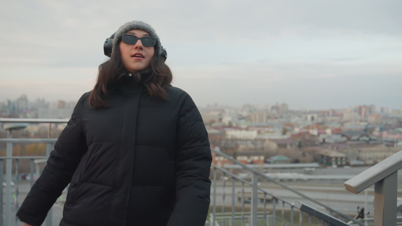Donna cammina lungo la ringhiera di un tetto, si ferma per ammirare il paesaggio urbano con le cuffie, un dondolio casuale e una postura riflessiva, la luce invernale e l'ampia vista urbana creano un'atmosfera cinematografica.