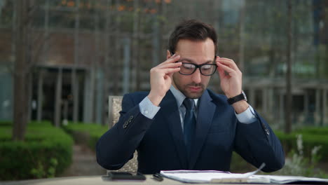 Successful-professional-bearded-man-in-business-suit-puts-on-glasses-and-reviews-paperwork-or-documents,-making-notes-sitting-in-a-park-outside-office-building---slow-dolly-in