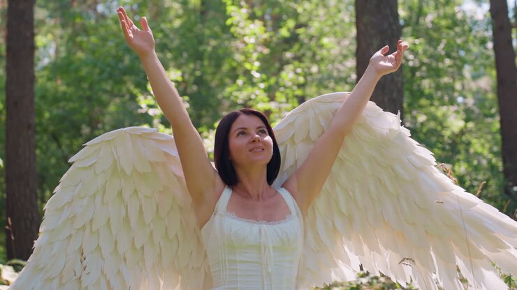 Fairy white goddess with grand angel wings walking gracefully through green woodland lifting arms toward bright sunlight, surrounded by glowing leaves and soft breeze
