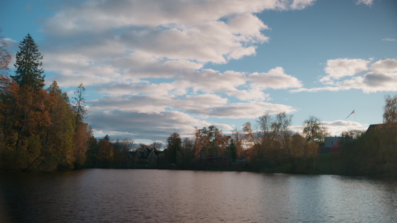 Wide shot of a lake with calm water and the sky has some clouds and the trees has orange leaves